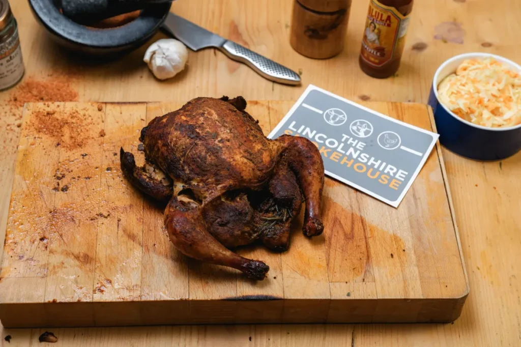 A charred whole chicken on a wooden cutting board next to seasoning bottles.