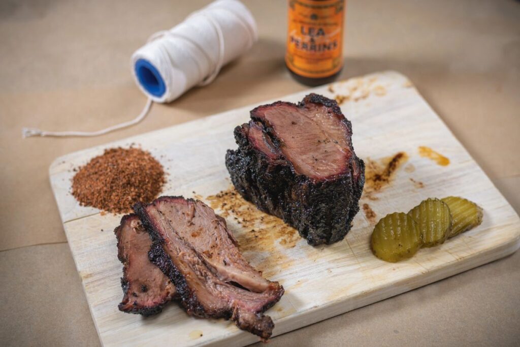 Sliced smoked brisket with spices and pickles on a wooden board.